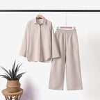 Tenue élégante beige composée d'une chemise à manches longues et d'un pantalon ample, suspendue sur un cintre, avec une plante en pot à côté.