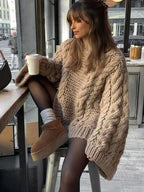 Femme en pull oversize tricoté beige, assise près d'une fenêtre, tenant un café. Mode automne-hiver, style décontracté, ambiance cosy.