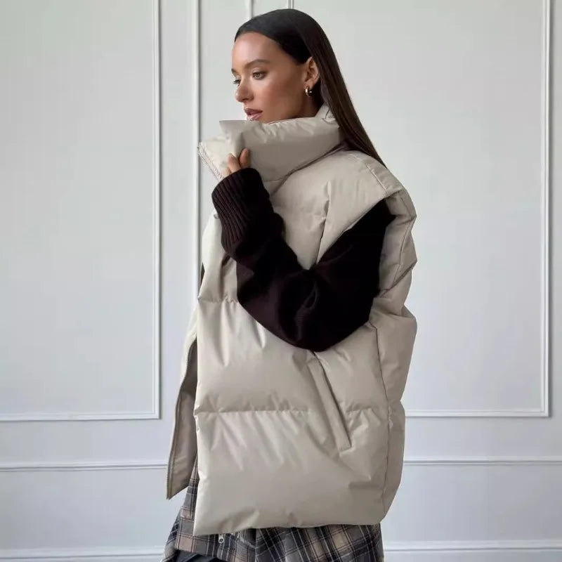 Femme portant une doudoune beige sans manches, pull noir, jupe à carreaux, posant de profil dans un intérieur moderne. Mode automne-hiver élégante.