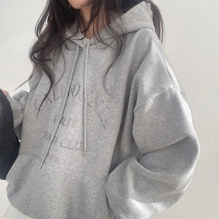 Femme portant un sweat à capuche gris avec inscription "California" en relief, style décontracté, mode automne, tendance vêtements confortables.