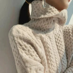 Femme portant un pull en laine torsadée beige avec col roulé, style automne-hiver, accessoirisé de boucles d'oreilles argentées.