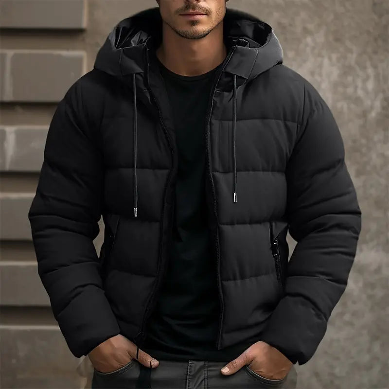 Homme portant une doudoune noire à capuche, style urbain, devant un mur en béton. Mode hiver, veste chaude, tendance masculine.