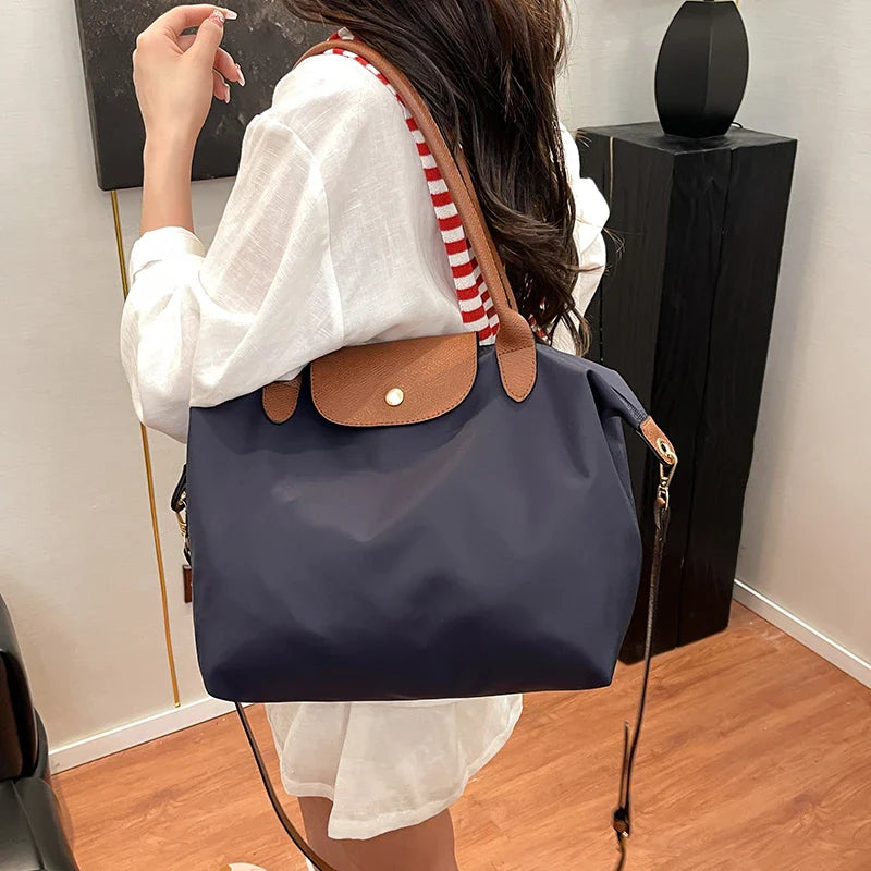 Femme portant un grand sac à main bleu marine avec détails en cuir marron, bandoulière rayée rouge et blanche, tenue décontractée, intérieur moderne.