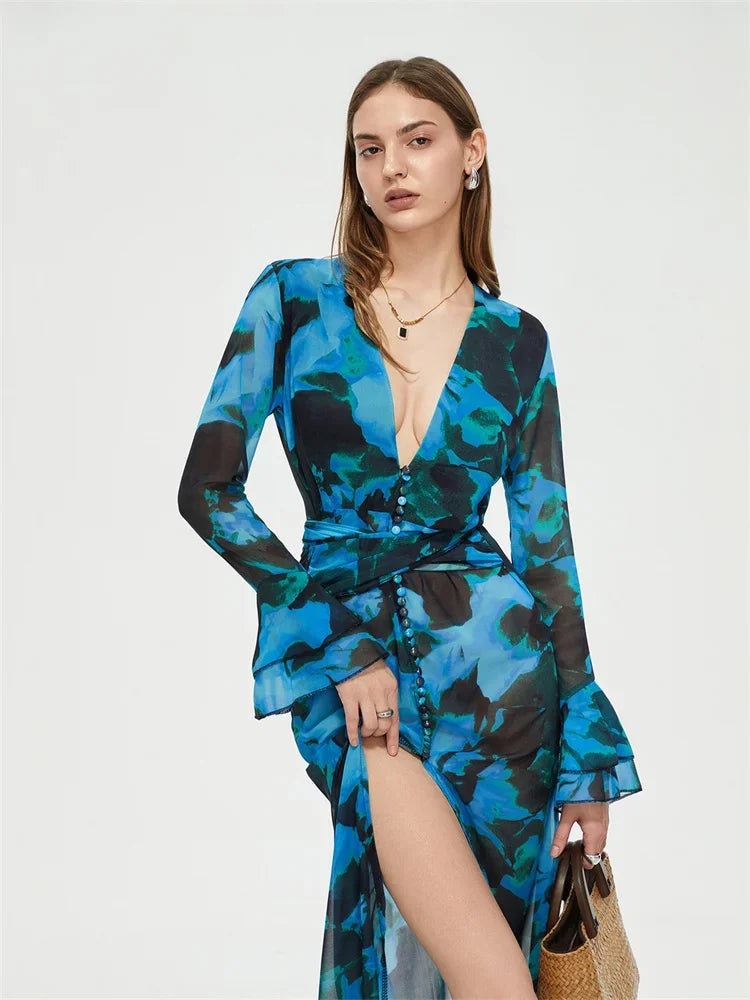 Robe longue bleue à motifs floraux, manches longues, décolleté plongeant, fente haute, portée par une femme tenant un sac en osier.