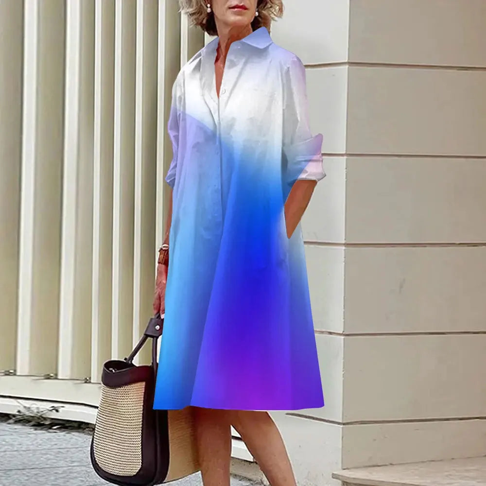 Robe dégradée bleu-violet, manches longues, portée par une femme avec sac à main beige et noir, devant un mur moderne. Mode élégante et tendance.