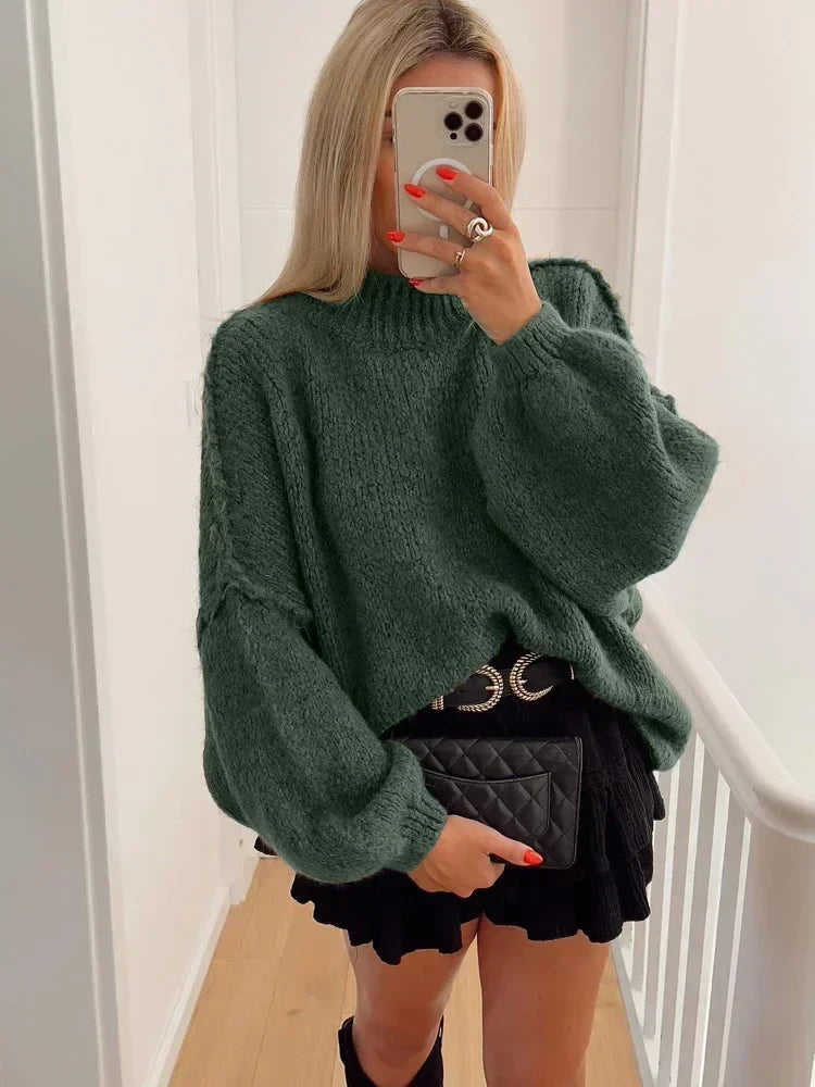 Femme en pull vert ample, jupe noire plissée, tenant un sac à main matelassé noir, prenant un selfie avec un iPhone dans un couloir lumineux.