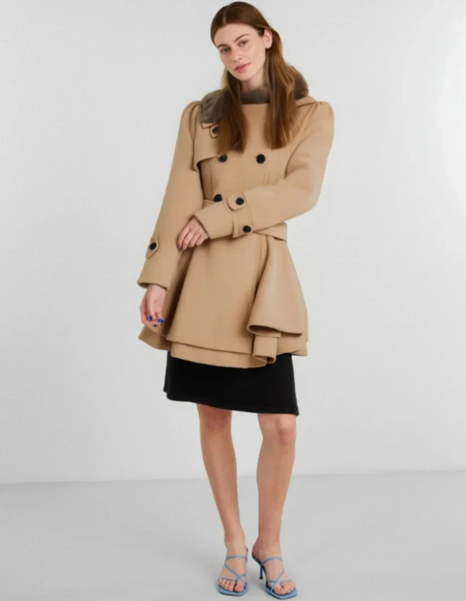 Femme portant un manteau beige élégant avec boutons noirs, jupe noire et sandales bleues. Mode automne-hiver, style chic et moderne.