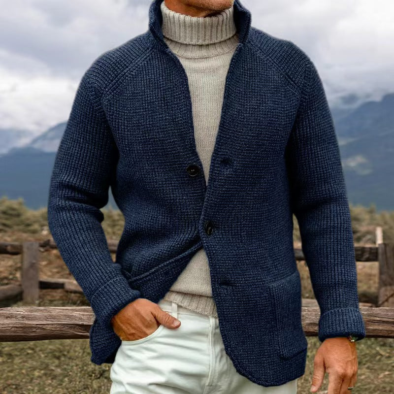 Homme portant un cardigan bleu en laine avec col roulé beige, debout devant un paysage montagneux. Mode masculine, tenue automne-hiver élégante.