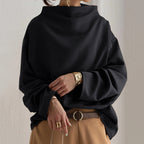 Femme portant un pull noir ample à col montant, manches longues, avec pantalon beige et ceinture marron. Mode automne-hiver élégante et décontractée.