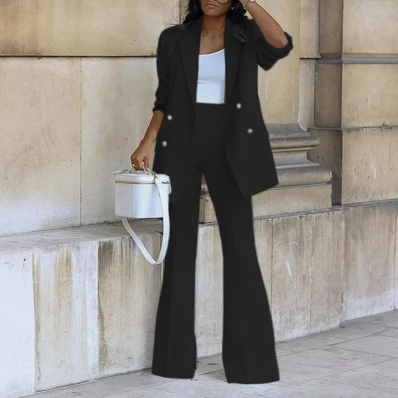 Femme en tailleur noir élégant, portant un sac blanc, posant contre un mur en pierre. Mode chic, tenue professionnelle, style urbain.