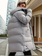 Homme portant une doudoune grise épaisse, debout dans un environnement urbain moderne. Mode hiver, manteau chaud, style urbain élégant.