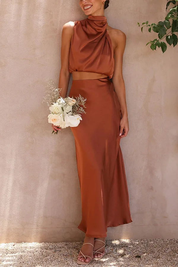 Robe longue satinée marron, sans manches, avec col drapé, portée par une femme tenant un bouquet de fleurs blanches et roses, sandales à talons.