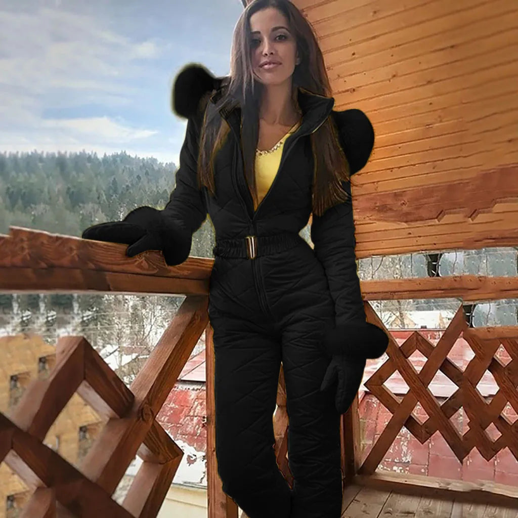 Femme en combinaison de ski noire sur un balcon en bois, forêt enneigée en arrière-plan. Mode hiver, tenue de ski élégante, paysage alpin.