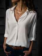 Femme portant une chemise blanche élégante, col ouvert, avec colliers dorés superposés, ceinture marron, fond sombre. Mode chic et décontractée.