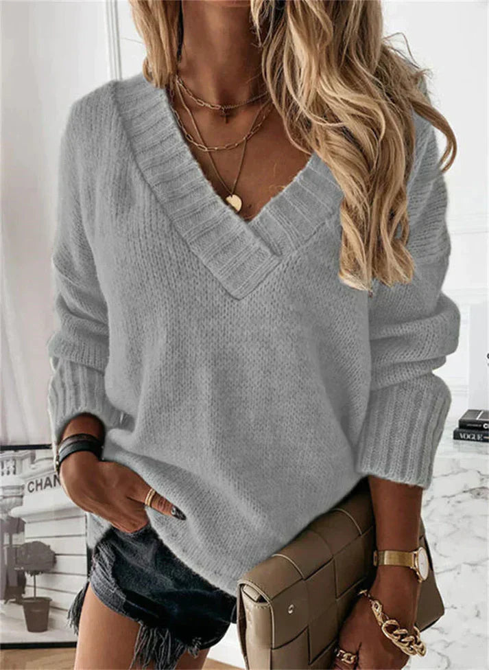 Femme portant un pull gris à col en V, short noir, sac à main beige, cheveux blonds ondulés, accessoires dorés. Mode automne chic et décontractée.
