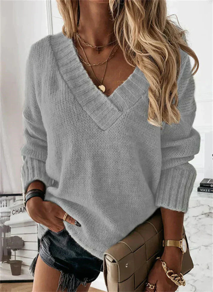 Femme portant un pull gris en tricot à col en V, avec des bijoux dorés et un sac à main marron, posant dans un intérieur moderne.