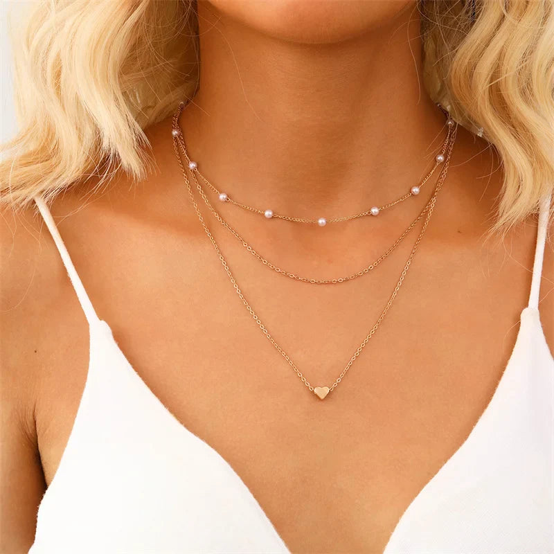 Collier multi-rangs en or avec perles et pendentif cœur, porté par une femme en débardeur blanc. Bijoux élégants, tendance mode féminine.