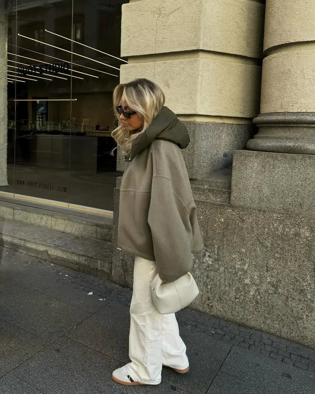 Femme en manteau oversize beige, pantalon blanc, sac à main blanc, lunettes de soleil, marchant en ville devant un bâtiment moderne.