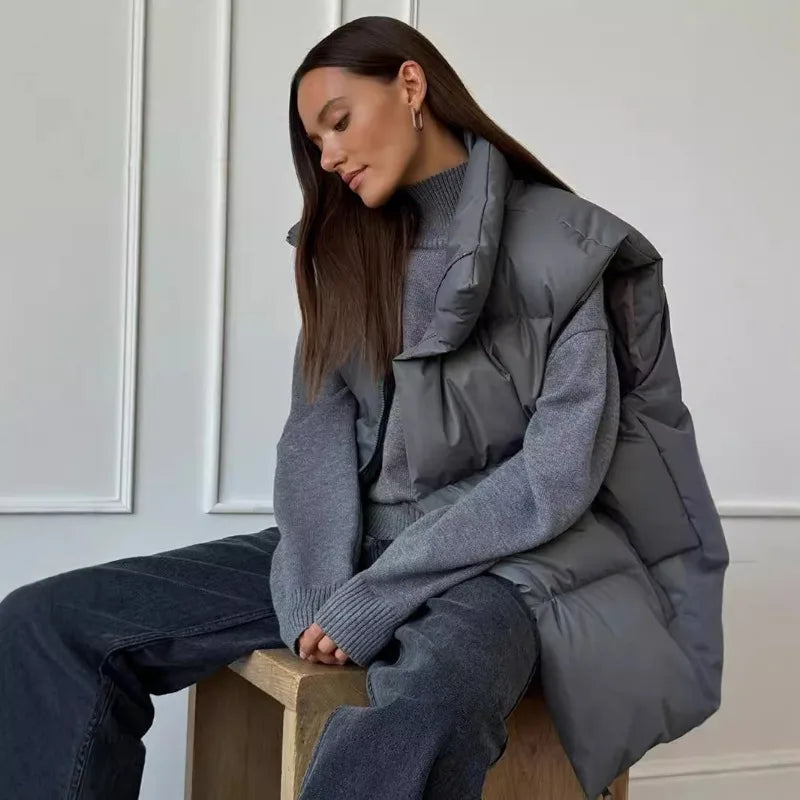 Femme portant un gilet matelassé gris sur un pull à col roulé, assise sur un tabouret en bois. Mode automne-hiver, style décontracté et élégant.