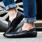 Chaussures mocassins en cuir noir pour homme, portées avec un jean bleu retroussé, devant une roue de voiture. Mode masculine élégante et décontractée.