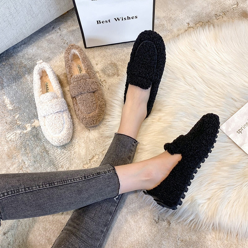Femme portant des mocassins noirs en peluche, avec deux paires supplémentaires en beige et marron sur un tapis. Chaussures confortables et tendance.