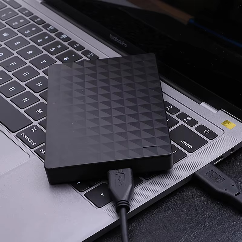 Disque dur externe noir connecté à un ordinateur portable via USB, posé sur un clavier d'ordinateur portable. Stockage de données portable et sécurisé.