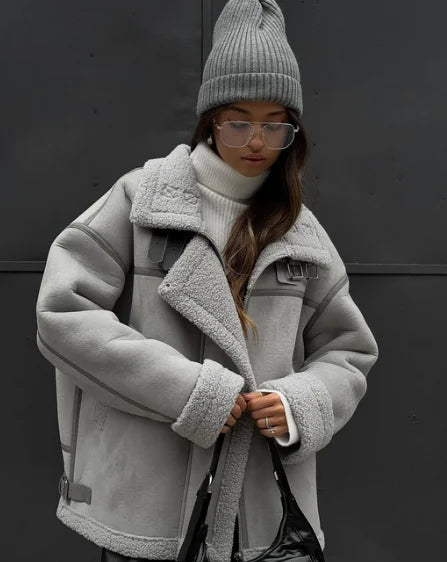 Femme portant un manteau en laine grise, pull col roulé blanc et bonnet assorti, style hiver urbain, mode tendance, lunettes transparentes.