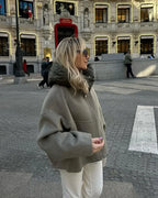 Femme en manteau gris et lunettes de soleil, debout devant un bâtiment historique illuminé, rue animée de Madrid. Mode hiver, style urbain.