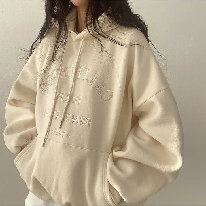 Femme portant un sweat à capuche beige avec inscription "California" en relief, style décontracté, mode automne-hiver, tendance vêtements confortables.