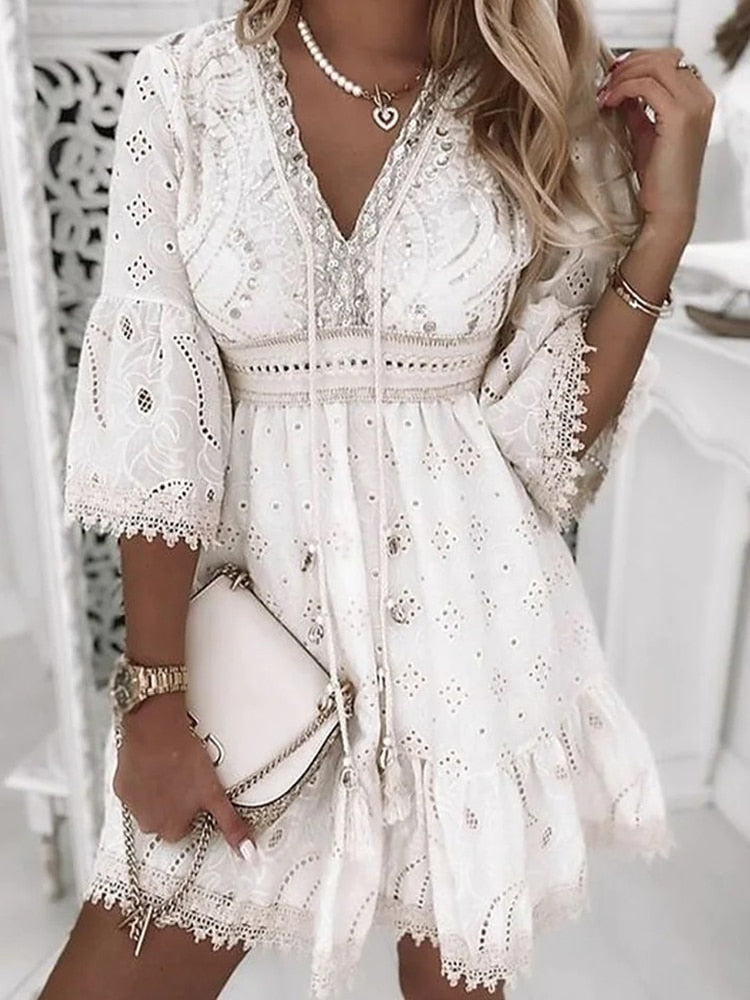 Robe blanche bohème chic en dentelle avec manches évasées, portée par une femme blonde. Accessoires: sac à main beige, collier perles.