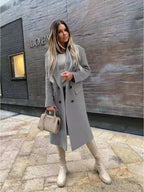 Femme élégante en manteau gris, portant un sac Prada beige, devant un mur moderne. Mode automne, style urbain chic, tendance 2023.