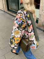 Femme portant une veste matelassée à motifs floraux colorés, écharpe verte, sac à main olive, marchant en ville. Mode automne, style urbain.