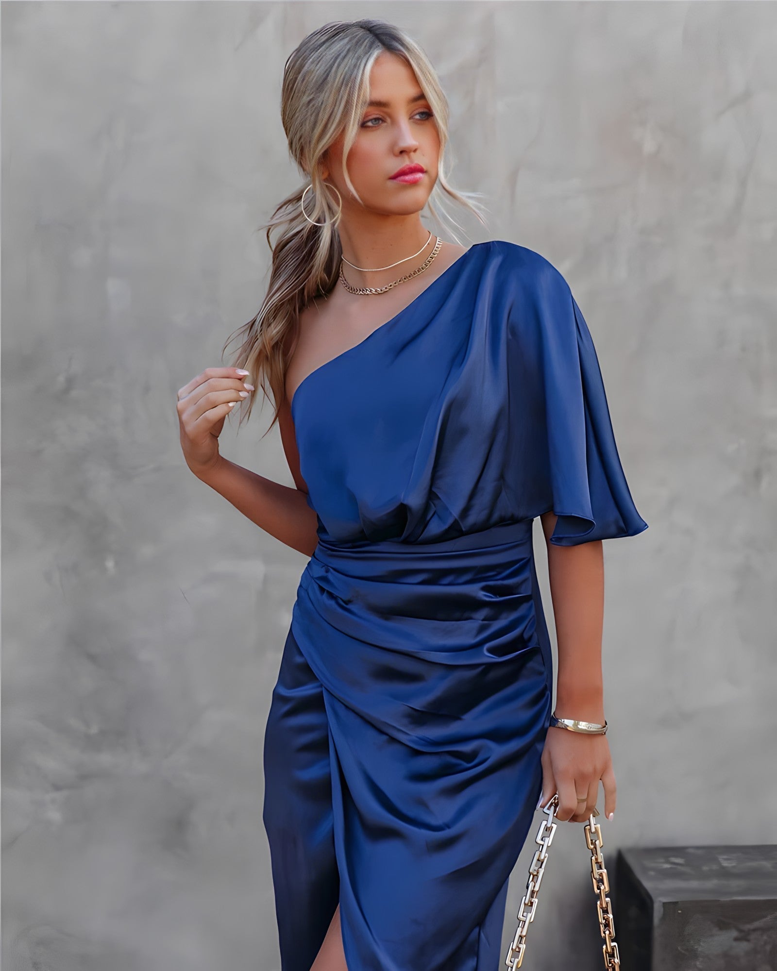 Femme en robe bleue satinée asymétrique, tenant un sac à chaîne dorée, posant devant un mur gris. Mode élégante, style chic, soirée glamour.