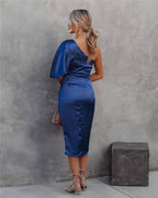Femme en robe bleue satinée asymétrique, cheveux blonds attachés, posant contre un mur gris. Mode élégante, tenue de soirée chic.