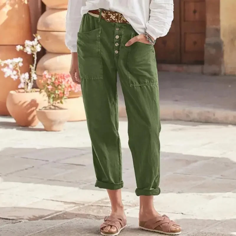 Personne portant un pantalon vert kaki taille haute avec boutons, ceinture marron, chemise blanche, sandales beiges, dans une rue pavée ensoleillée.