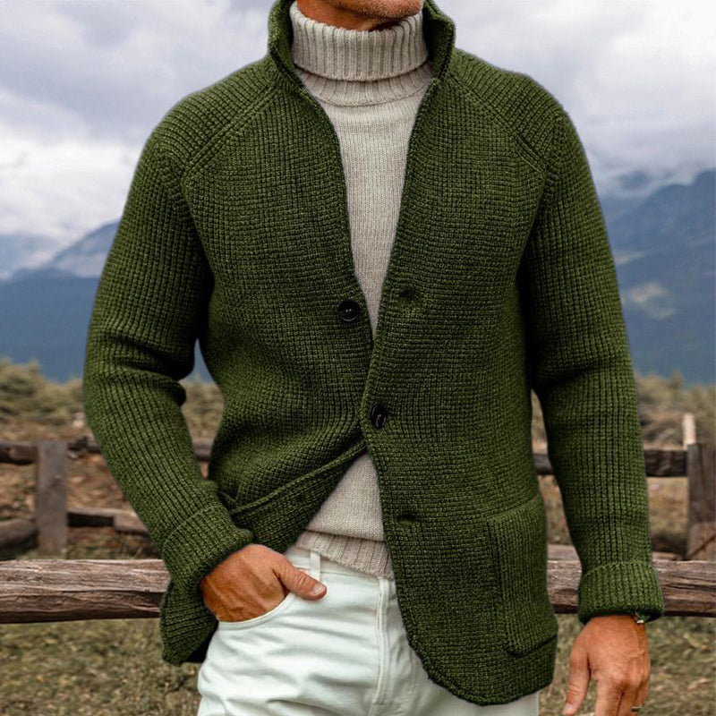 Homme portant un cardigan vert en laine avec col montant, sur un pull beige, devant un paysage montagneux. Mode masculine, style automne-hiver.