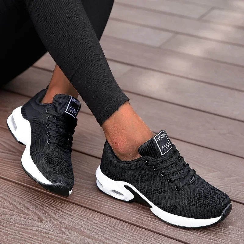 Chaussures de sport noires pour femme, design moderne, semelle blanche, confortables, idéales pour la course et le fitness, portées sur terrasse en bois.