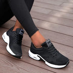 Chaussures de sport noires pour femme, design moderne, semelle blanche, confortables, idéales pour la course et le fitness, portées sur terrasse en bois.