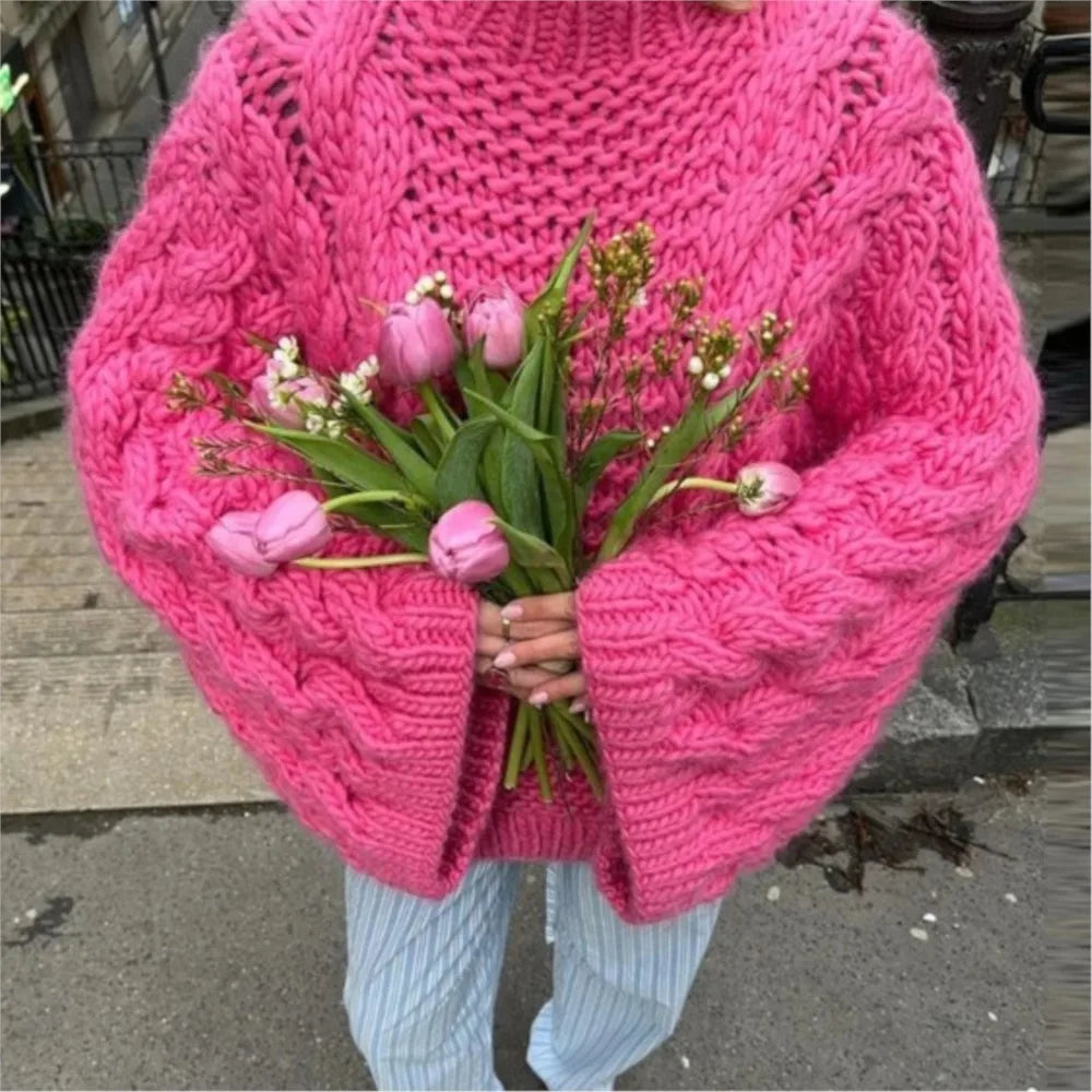 Personne portant un pull rose en tricot épais, tenant un bouquet de tulipes roses et de muguet, debout sur un trottoir urbain.