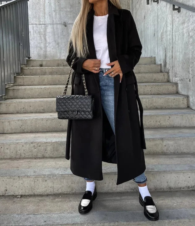 Femme en manteau noir, jean bleu, sac à main matelassé, escaliers en béton, mode urbaine chic, style automne, chaussures mocassins noires.