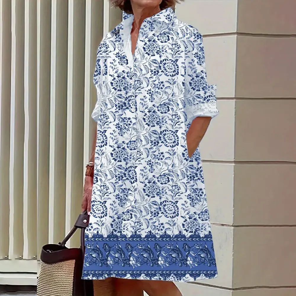 Robe d'été femme à motifs floraux bleus et blancs, manches retroussées, col en V, devant un mur beige, sac à main en osier à la main.