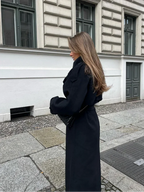 Femme en manteau noir élégant, cheveux longs, marchant sur un trottoir pavé devant un bâtiment classique. Mode automne chic, style urbain.