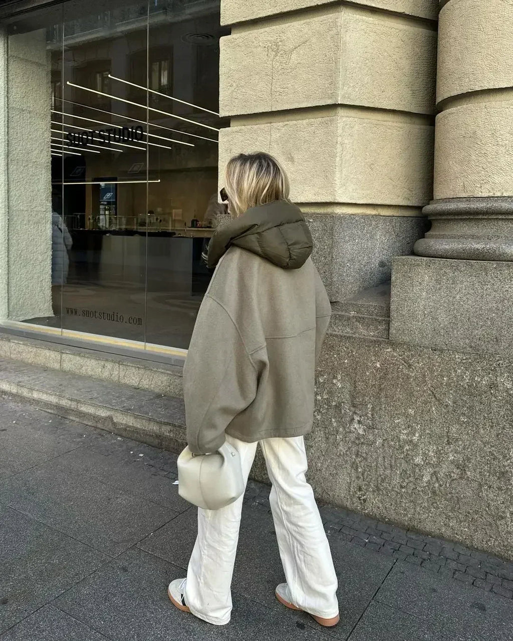 Femme en manteau oversize gris et pantalon blanc, portant un sac à main beige, marche devant une vitrine moderne en ville. Mode urbaine chic.