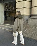 Femme en manteau oversize gris et pantalon blanc, portant un sac à main beige, marche devant une vitrine moderne en ville. Mode urbaine chic.