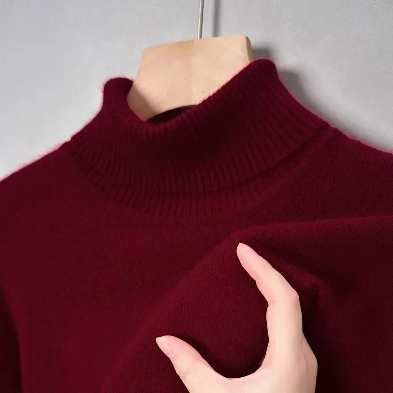 Pull-over col roulé bordeaux en laine, main touchant le tissu doux, mode automne-hiver, vêtement chaud et élégant, fond gris.