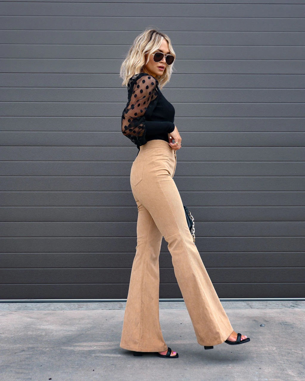 Femme en tenue chic: pantalon évasé beige, haut noir à manches transparentes à pois, lunettes de soleil, posant devant un mur gris. Mode élégante.