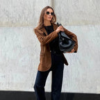 Femme élégante portant un blazer marron, lunettes de soleil et sac noir, posant devant un mur en marbre. Mode automne chic, style urbain.