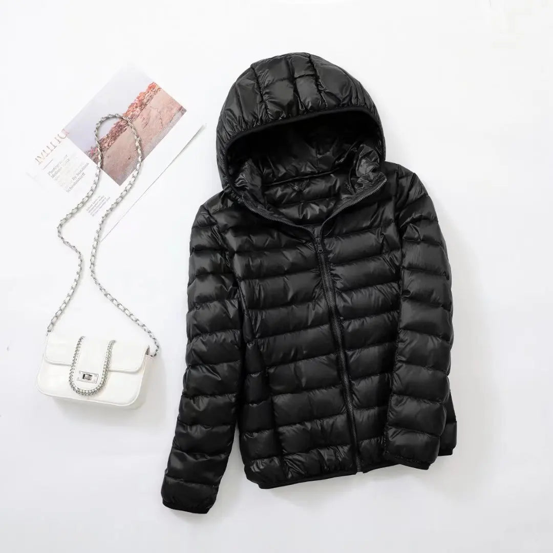 Veste doudoune noire à capuche posée à plat, accompagnée d'un sac à main blanc avec chaîne argentée, sur fond blanc. Mode hiver tendance.