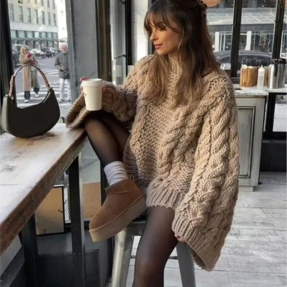 Jeune femme en pull oversize beige, collants noirs et bottes marron, assise dans un café moderne, tenant un gobelet de café. Mode hiver décontractée.