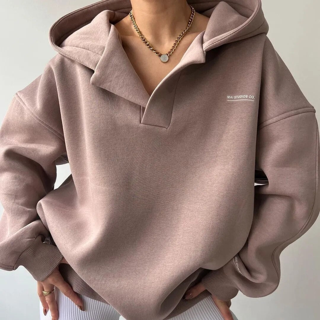Femme portant un sweat à capuche beige, col ouvert, chaîne dorée, style décontracté. Mode automne, tendance vêtements confortables.